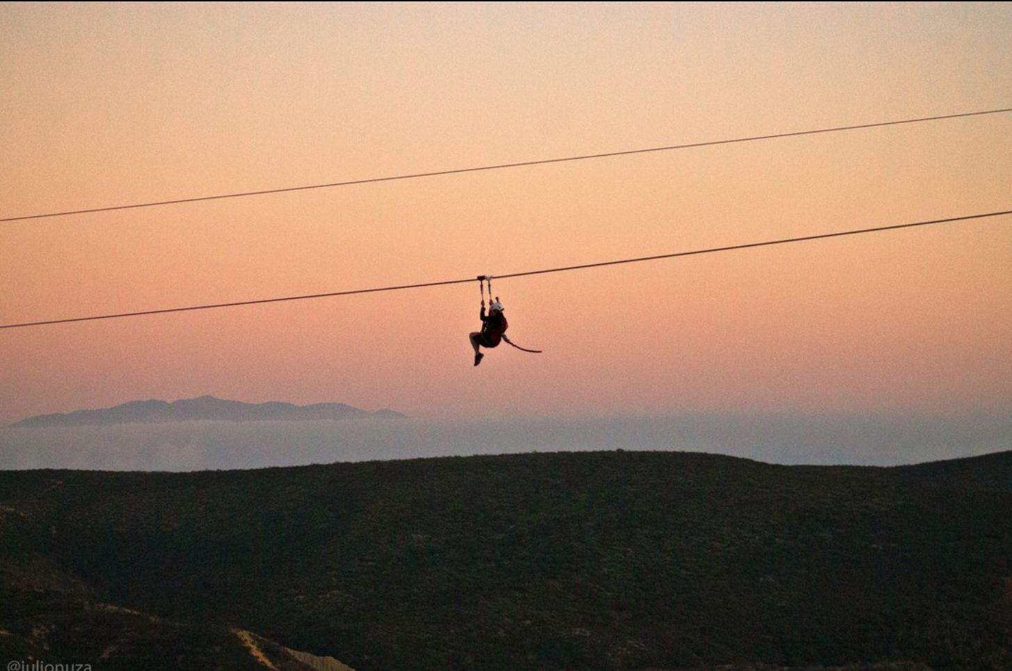 Una persona deslizándose en tirolesa al atardecer sobre colinas oscuras con un cielo de color naranja pastel.