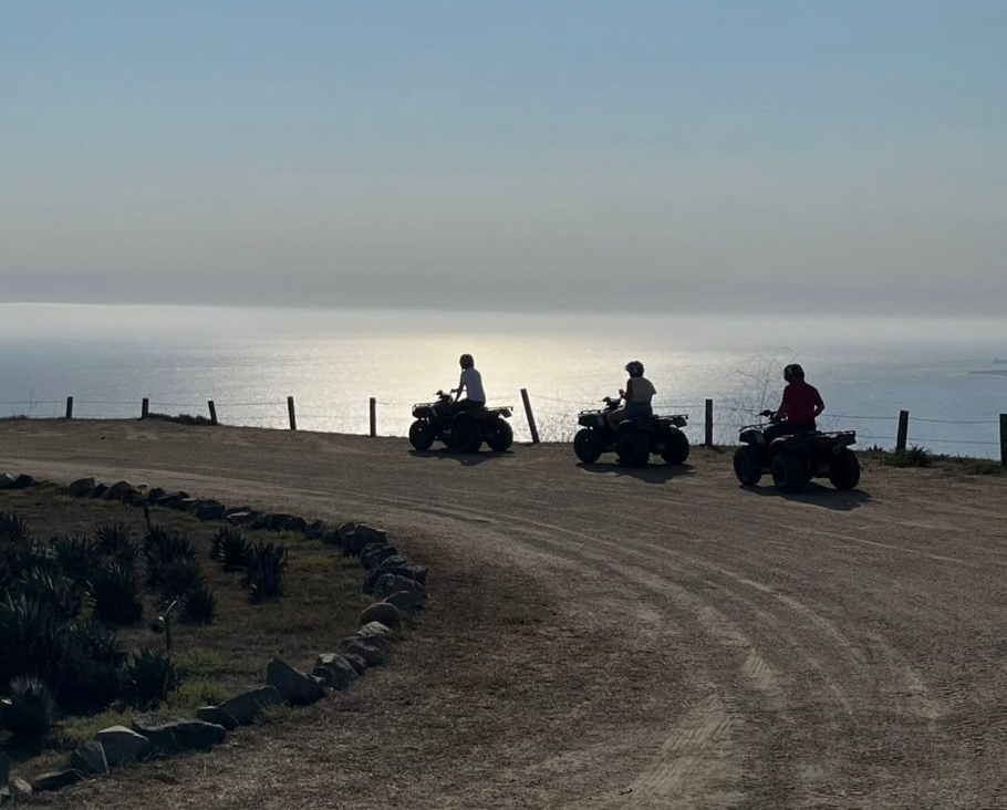 Tres personas en vehículos todoterreno en un camino de tierra costero al atardecer, con vistas al océano.