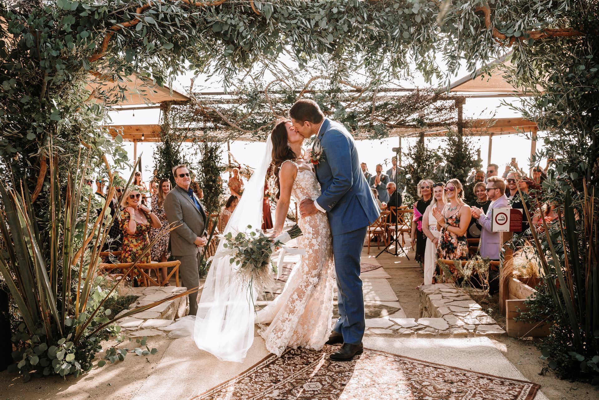Una pareja de recién casados se besa bajo un arco en una ceremonia en el jardín, con los invitados sentados cerca.