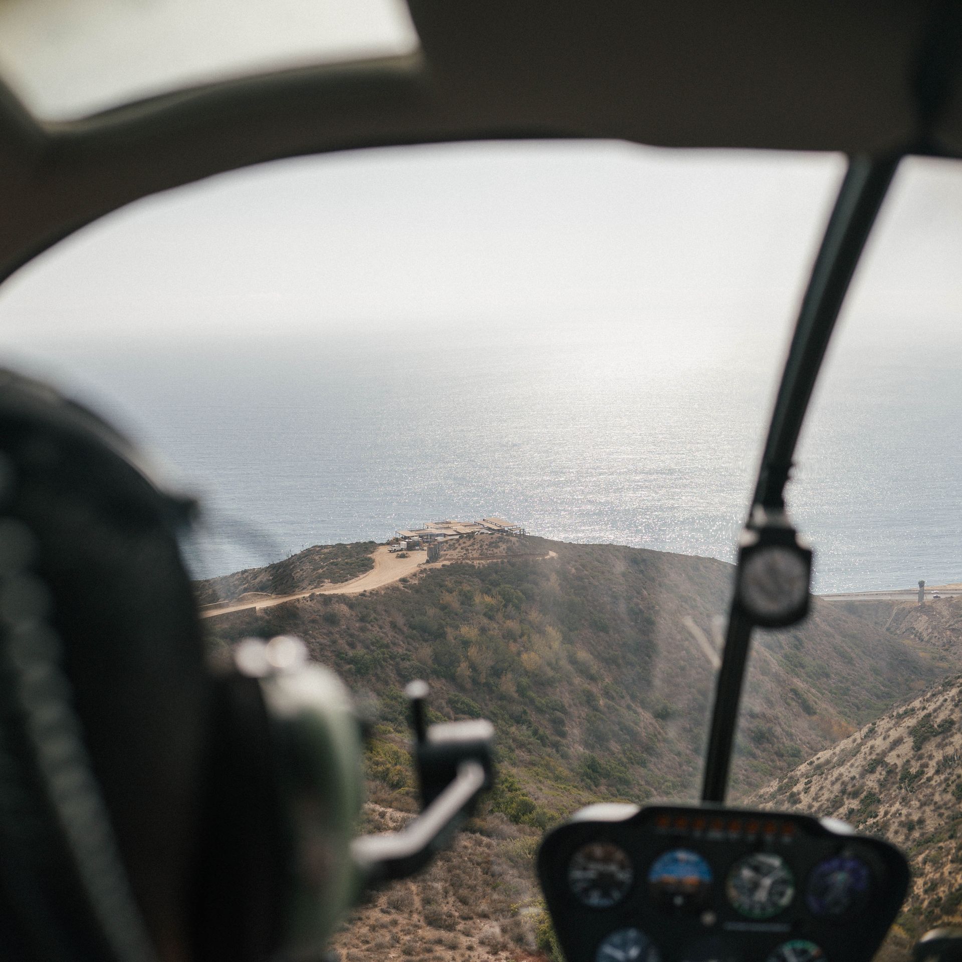 Vista desde la cabina de un helicóptero sobre una costa rocosa y el océano bajo un cielo brumoso.