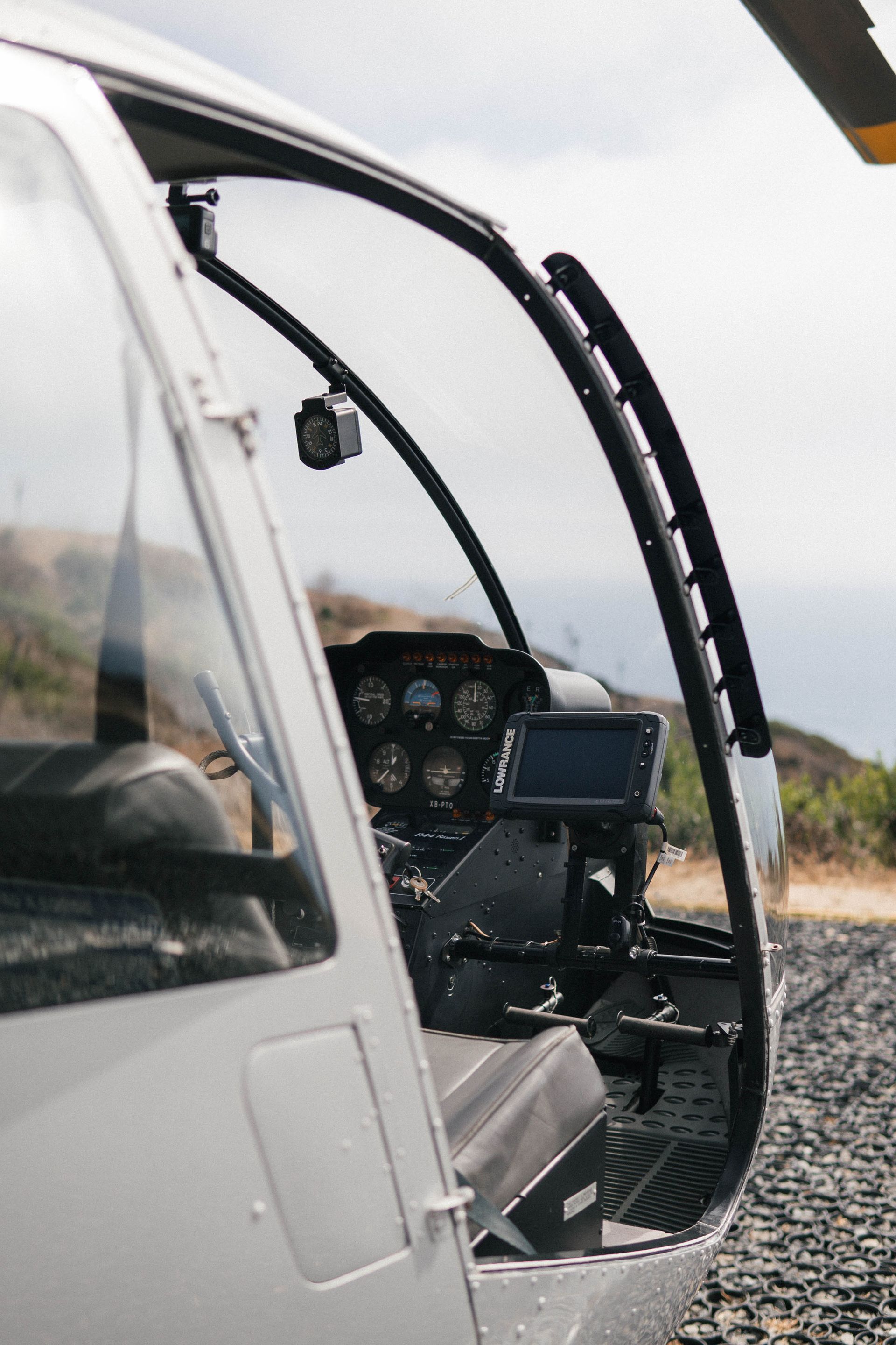 Cabina de helicóptero abierta con panel de instrumentos y techo transparente en una zona de aterrizaje rocosa al aire libre.
