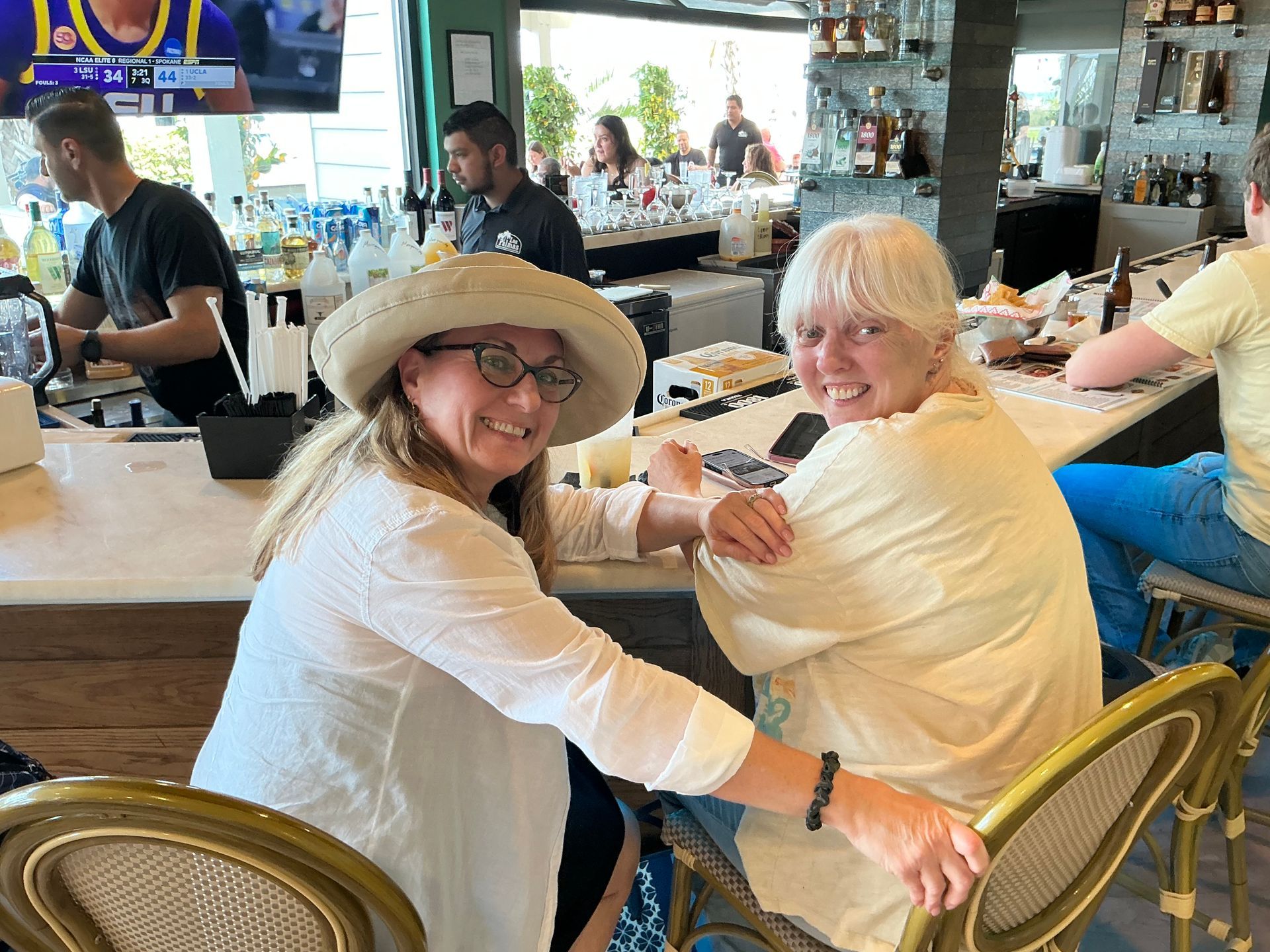 Two women smiling at a bar, one wearing a hat. Bar staff visible in the background.