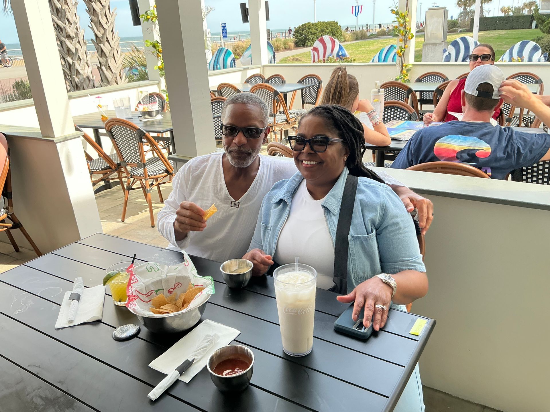 Couple smiles at outdoor table, holding drinks and food, with beach in the background.