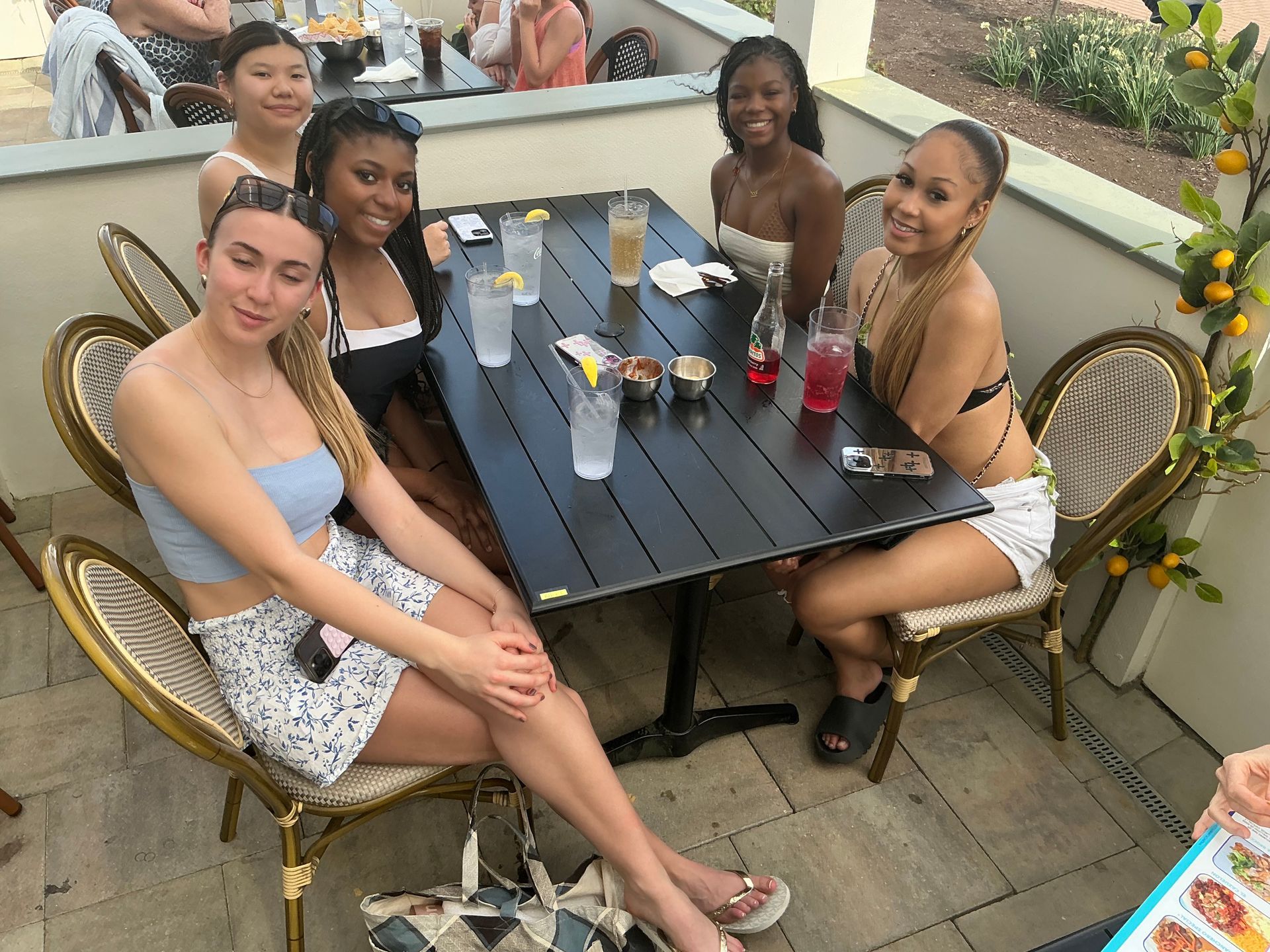 Six people at a table outdoors. Drinks on the table. Chairs are ornate. Sunny setting.