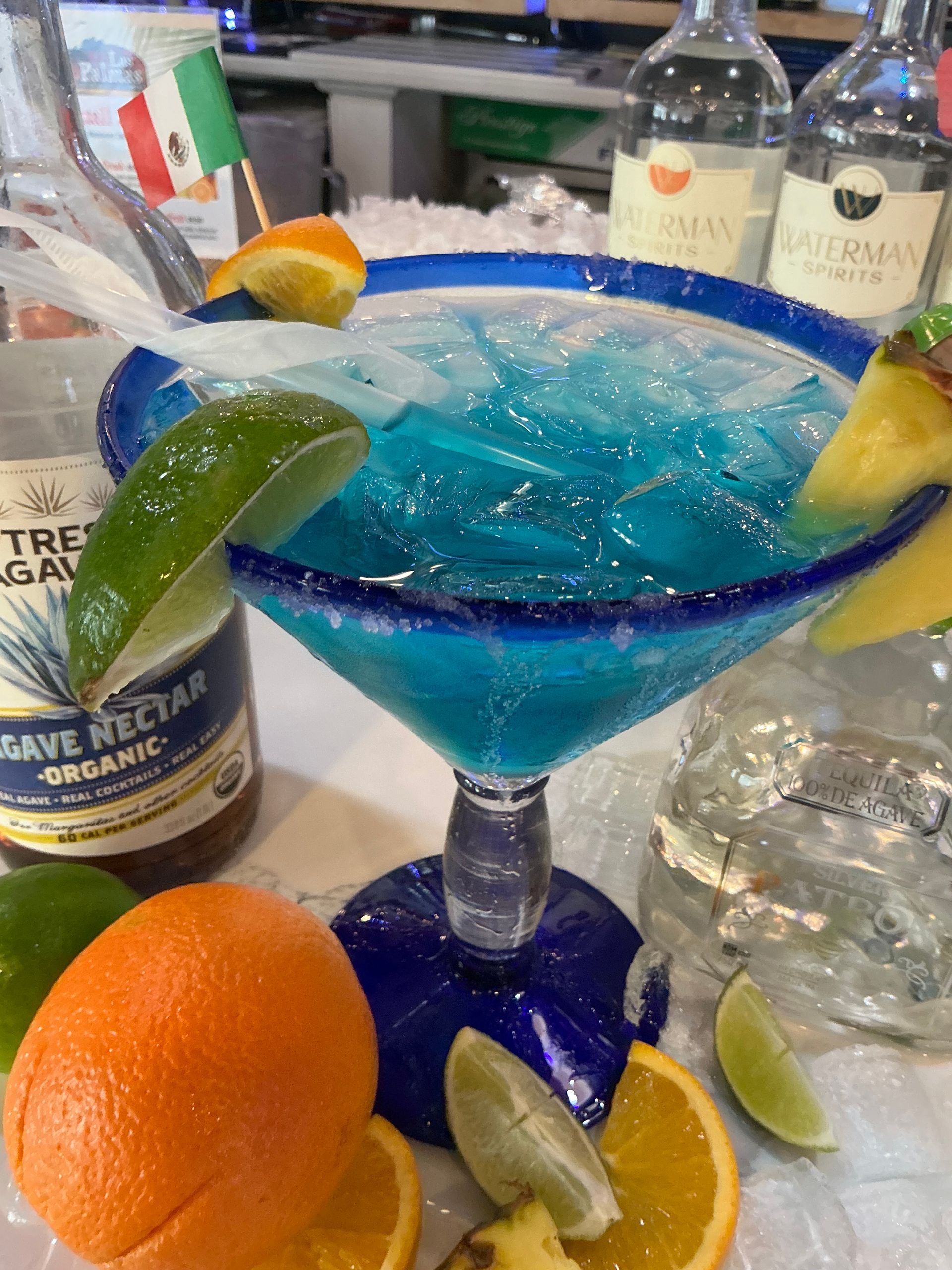 Blue margarita cocktail in a large glass with fruit garnishes and tequila bottles.