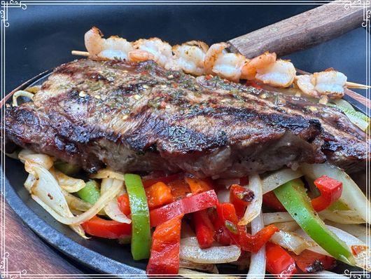 Sizzling steak and shrimp skewers atop sauteed peppers and onions on a hot skillet.