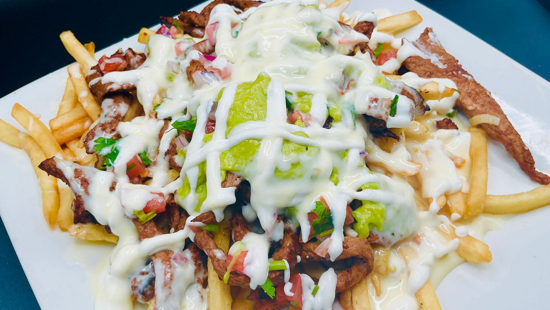 Fries topped with grilled meat, guacamole, salsa, and white sauce on a white plate.