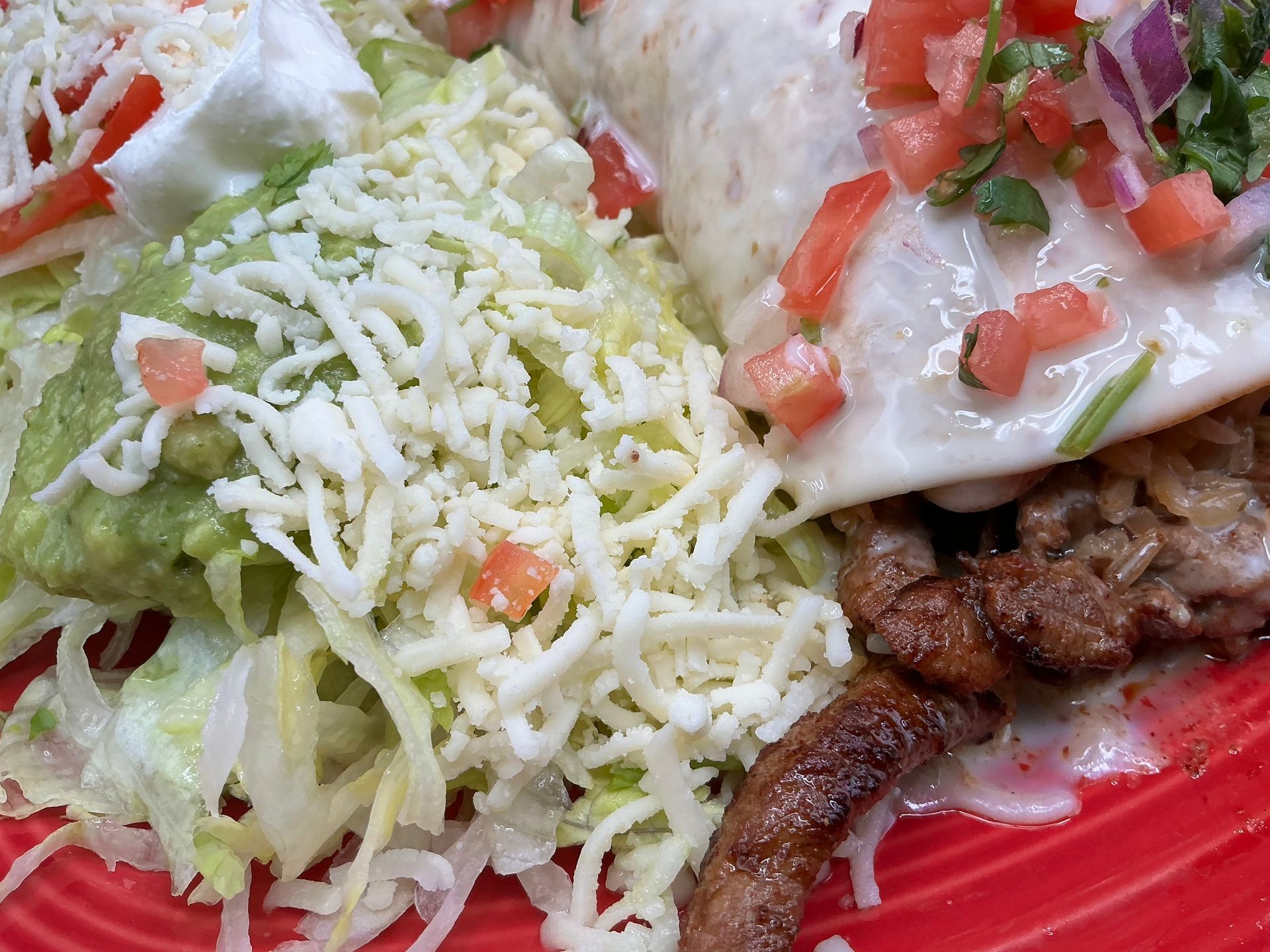Burrito, guacamole, salad, and meat on a red plate, topped with cheese and pico de gallo.