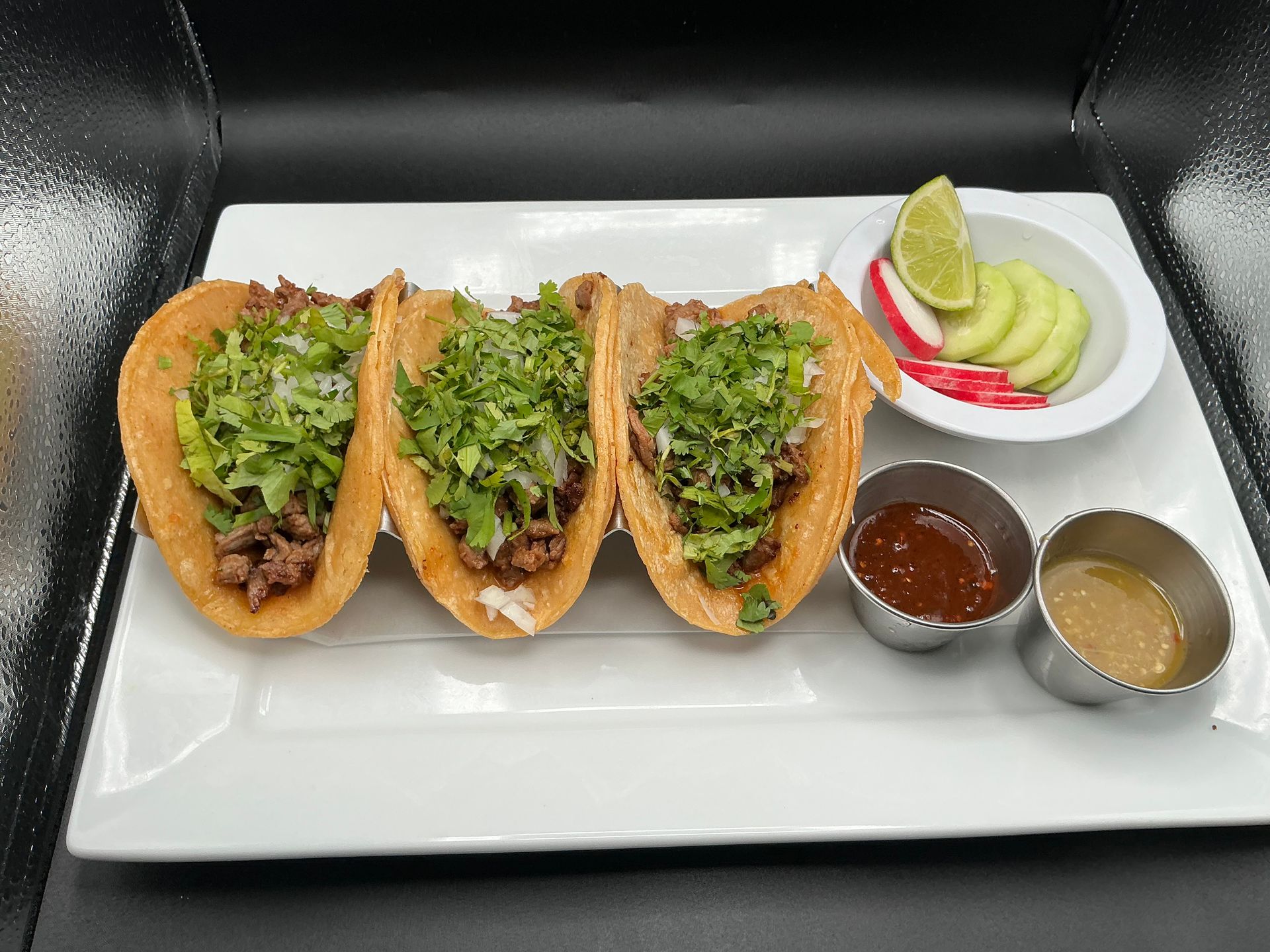 Three tacos with toppings on a white plate, served with sauces and sliced cucumbers.