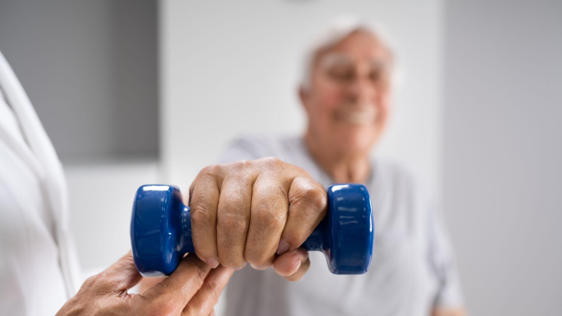 Person's hand gripping a blue dumbbell, guided by another hand, elderly person blurred in background.