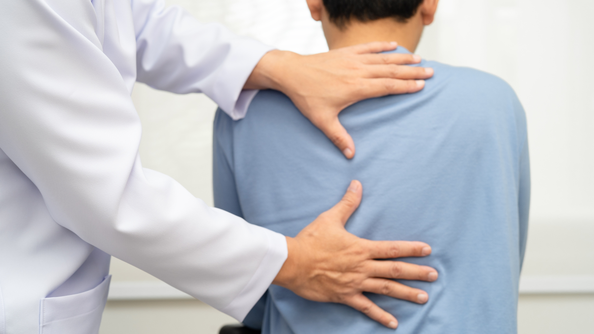 Doctor examines a patient's back, hands on back, white coat, blue shirt, indoors.