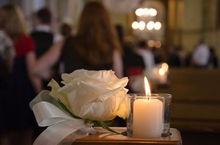 White rose beside a lit candle on a table, with a softly blurred indoor gathering in the background