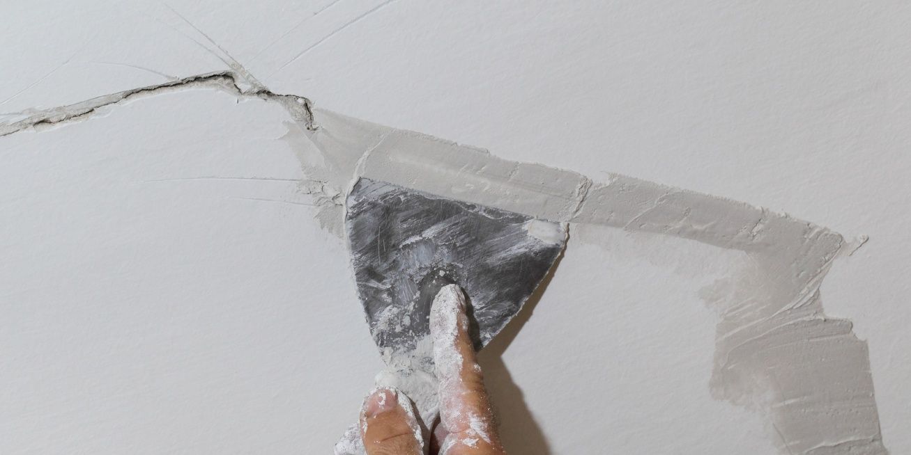 A hand using a putty knife to apply spackle to a crack on a white wall.