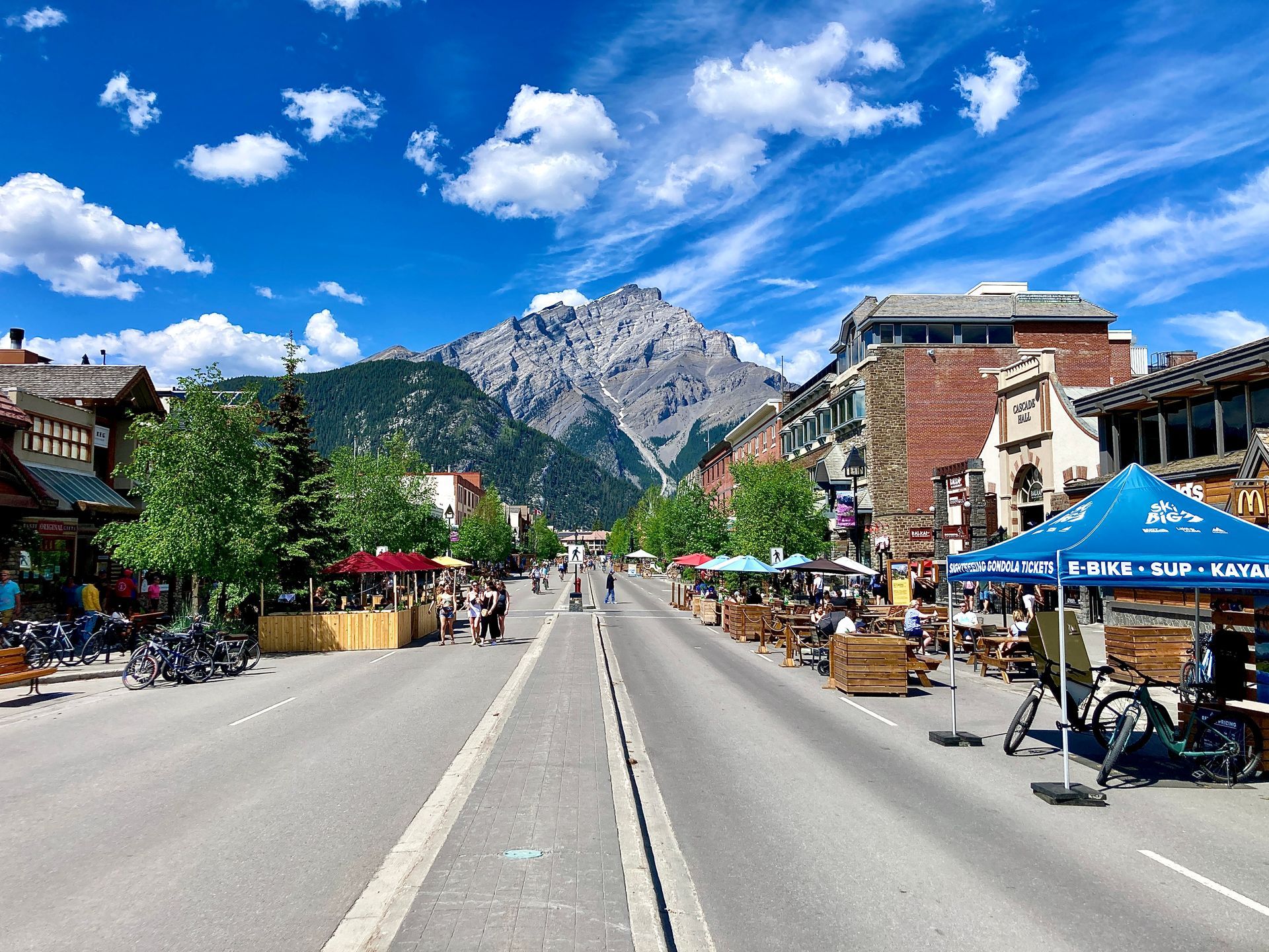 Mountain town street with shops and cyclists, leading to rocky peaks under a bright blue sky