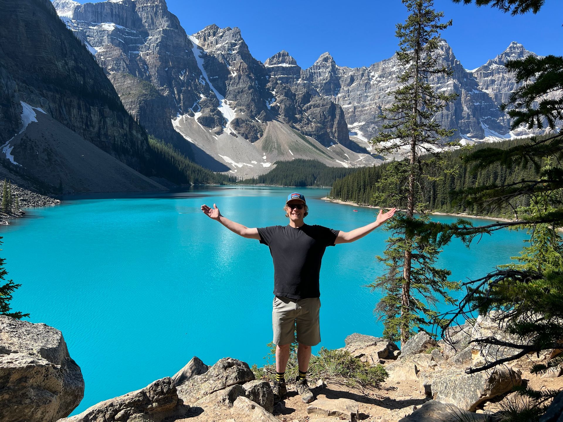 Person standing with arms outstretched by a turquoise alpine lake surrounded by mountains