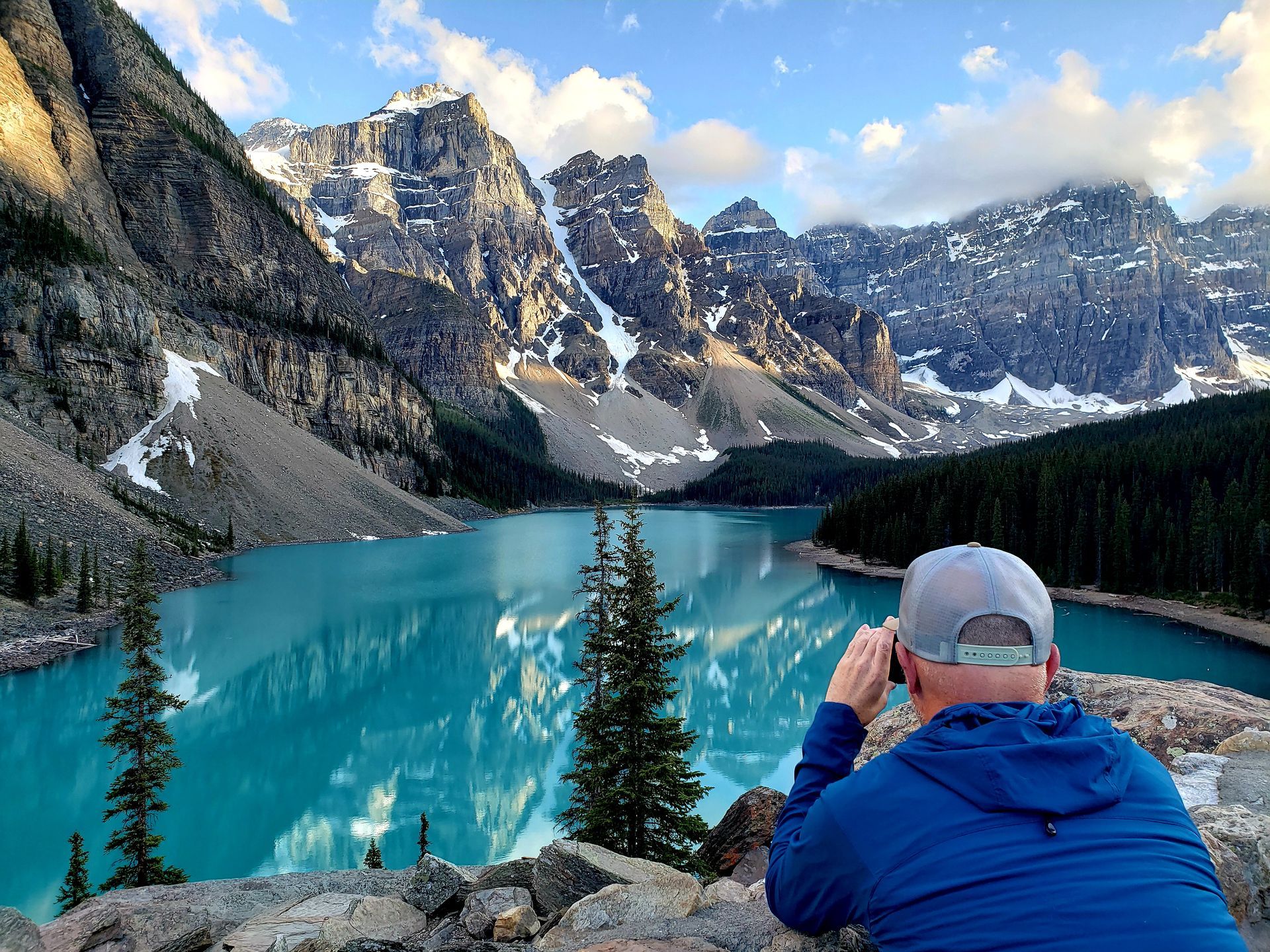 Person in blue jacket photographing a turquoise mountain lake with snowy peaks under clouds