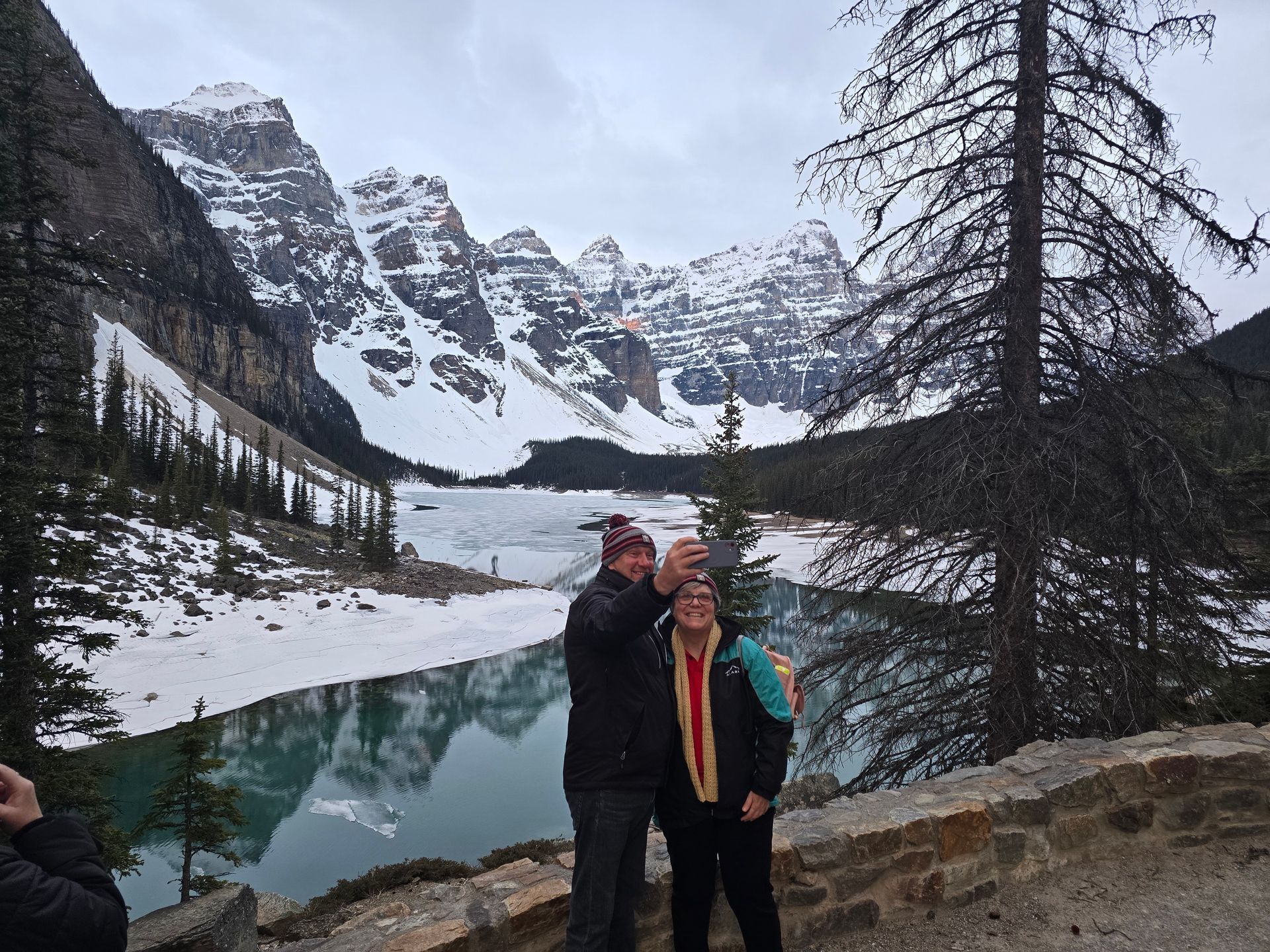 Two people posing by a turquoise alpine lake with snowy mountains in the background.