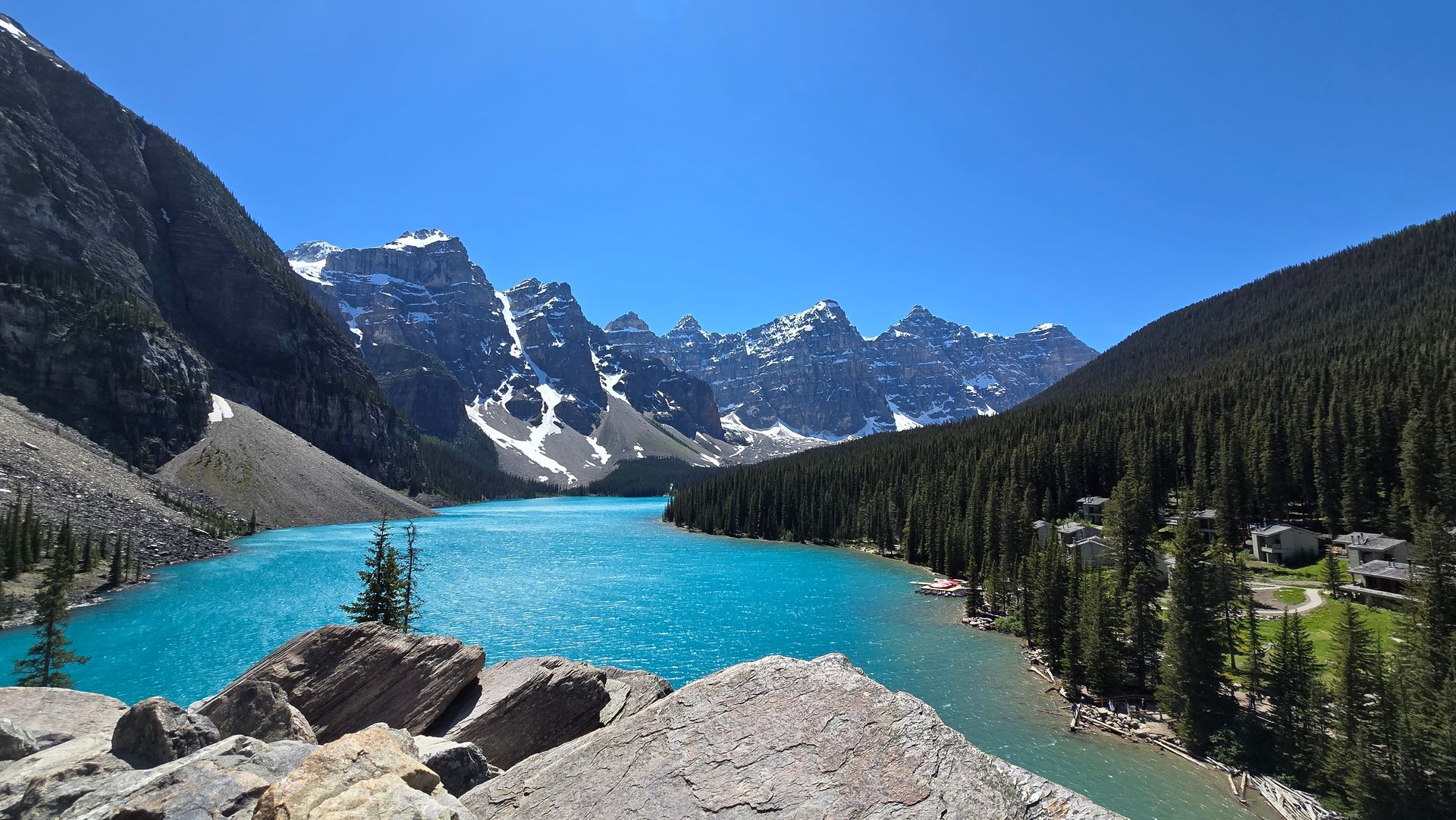 Turquoise alpine lake with rocky shore, pine forest, and snowcapped mountains under a clear blue sky