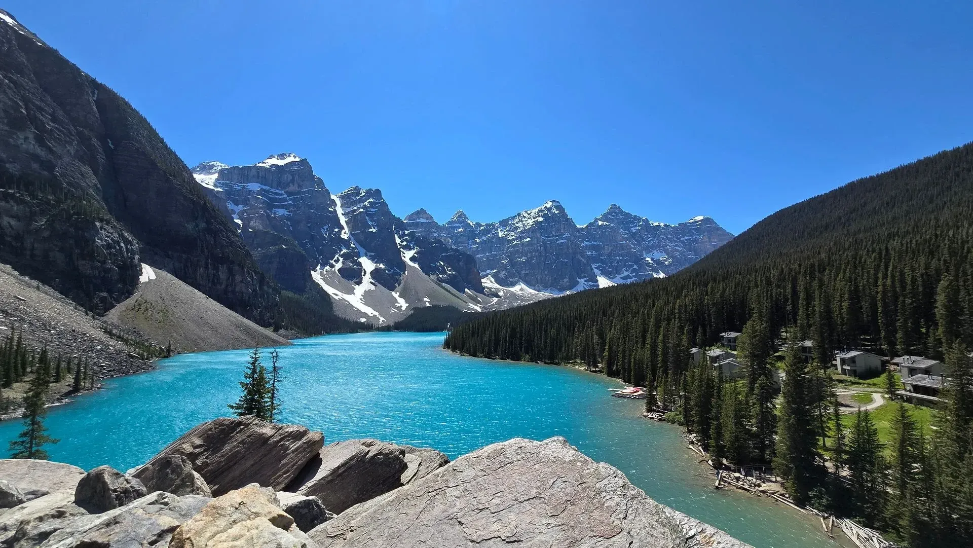 Turquoise alpine lake with rocky shore, pine forest, and snow-capped mountains under a clear blue sky