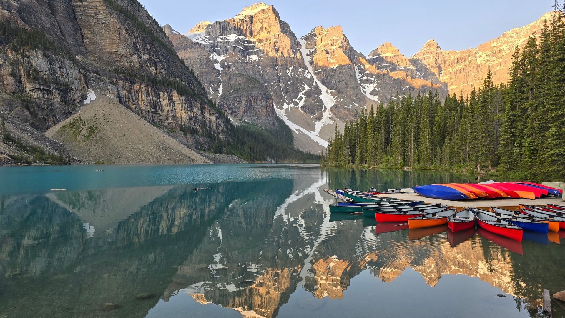Colorful kayaks lined up on a mountain lake with mirrored peaks and pine forest at sunrise