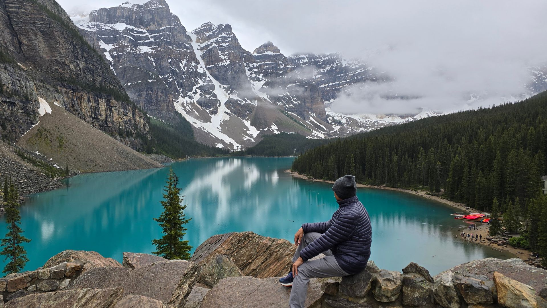 Person sitting on rocks overlooking a turquoise mountain lake and snowy peaks under cloudy skies