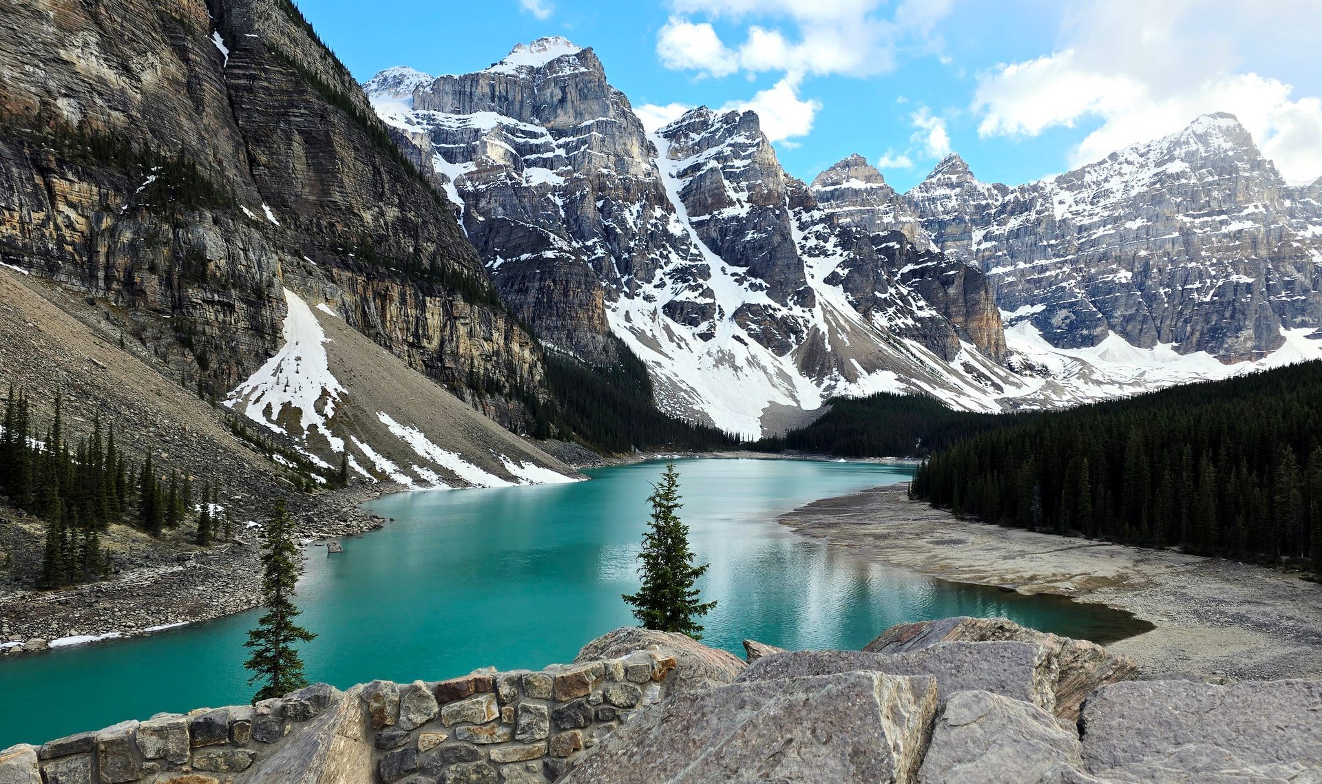 Turquoise alpine lake with snowcapped mountains, rocky shore, and evergreen trees under a blue sky.