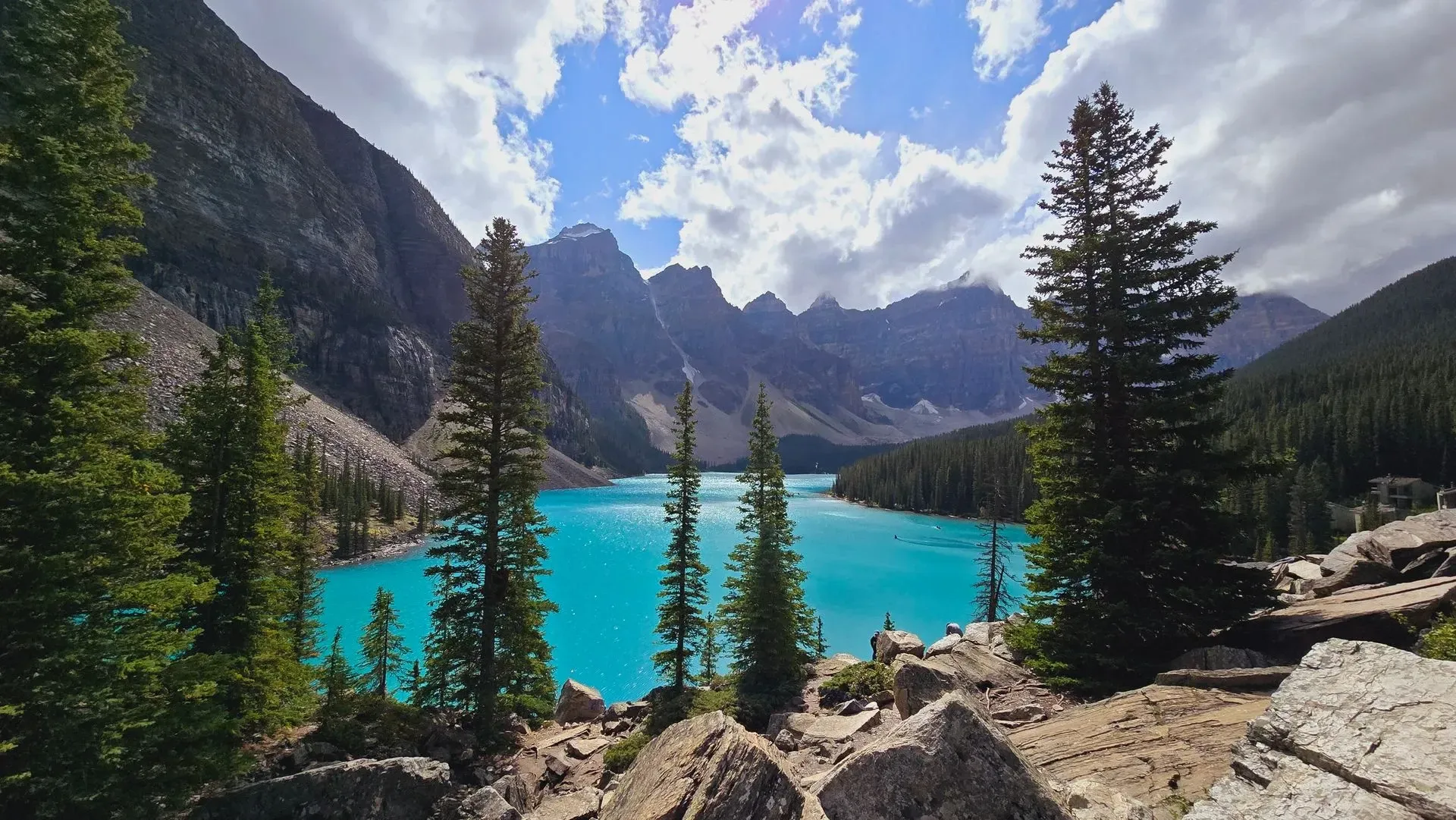 Moraine lake framed by pine trees and rocky peaks under a partly cloudy sky