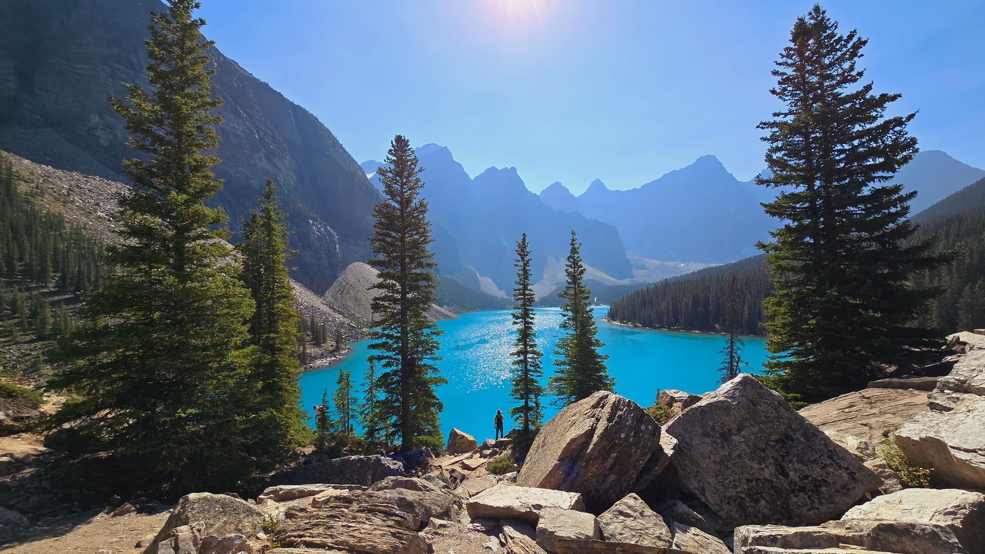 Turquoise alpine lake with pine trees, rocky shore, and sunlit mountain peaks under a clear sky