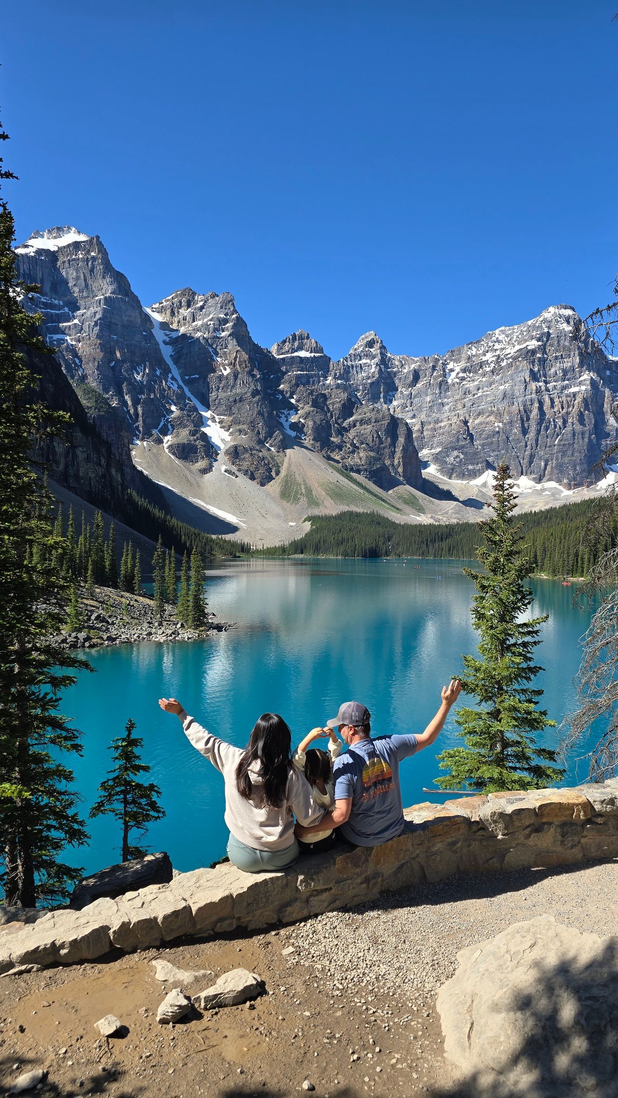 Two hikers on a rock overlook a turquoise alpine lake and snowy mountain peaks under a clear blue sky