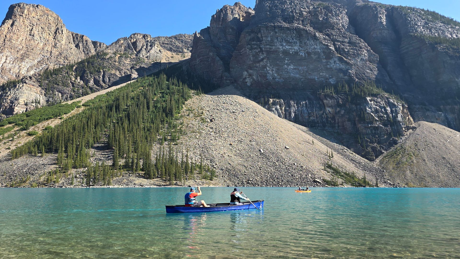 Two people in a blue canoe on a clear mountain lake with rocky cliffs and forested slopes behind them