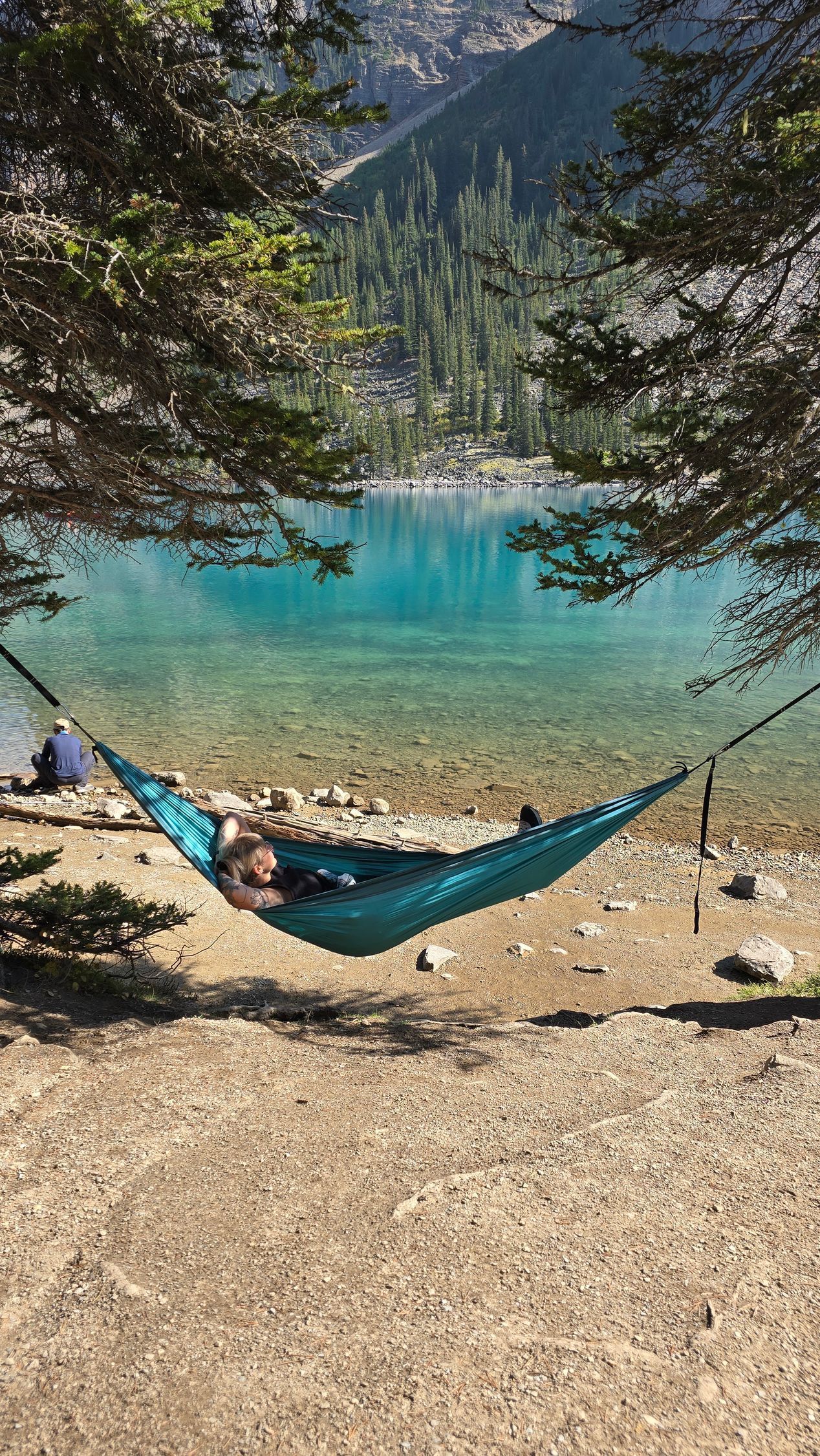 Blue hammock by a turquoise mountain lake, framed by trees and rocky shore.