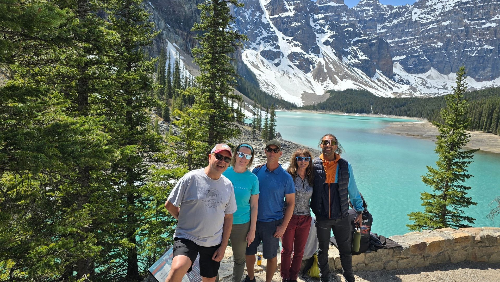 Group posing on a rocky trail beside a turquoise alpine lake with snow-covered mountains in the background