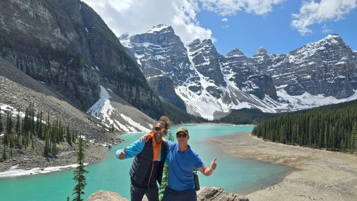 Two hikers posing by a turquoise mountain lake with snowy peaks and steep rocky cliffs.