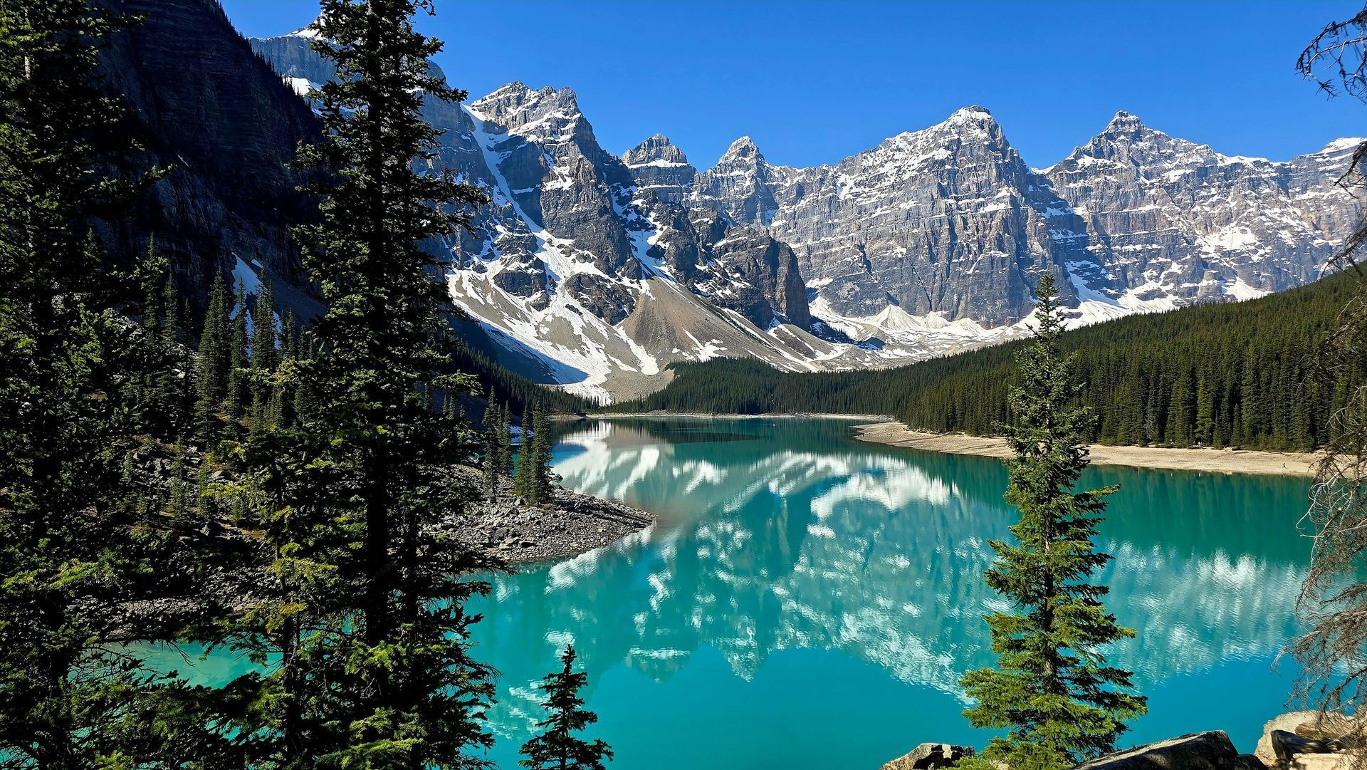 Turquoise mountain lake with pine trees and snow-capped peaks under a clear blue sky