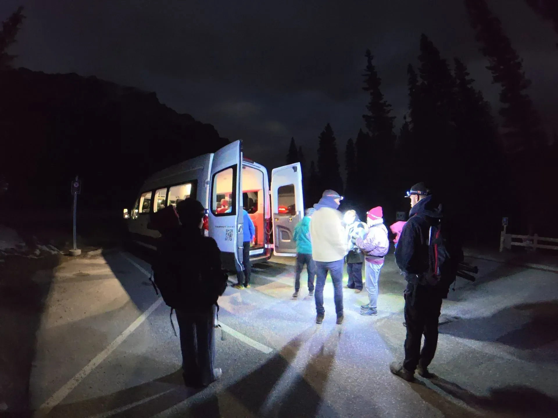 People boarding the shuttle at night on a dark road, lit by the bus and a flashlight.