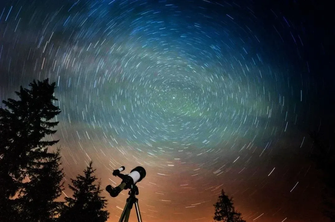 Silhouette of a telescope under a swirling starry night sky with silhouetted trees