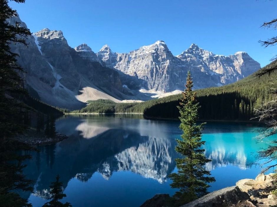 Mountain lake with turquoise water, pine forest, and jagged snowy peaks reflected in a calm mirror surface