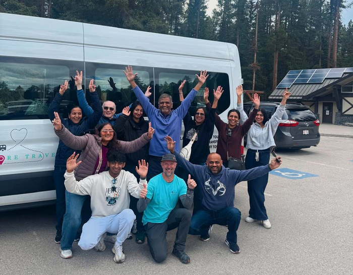 Group of people posing and waving in front of a white van in a parking lot with trees and buildings nearby