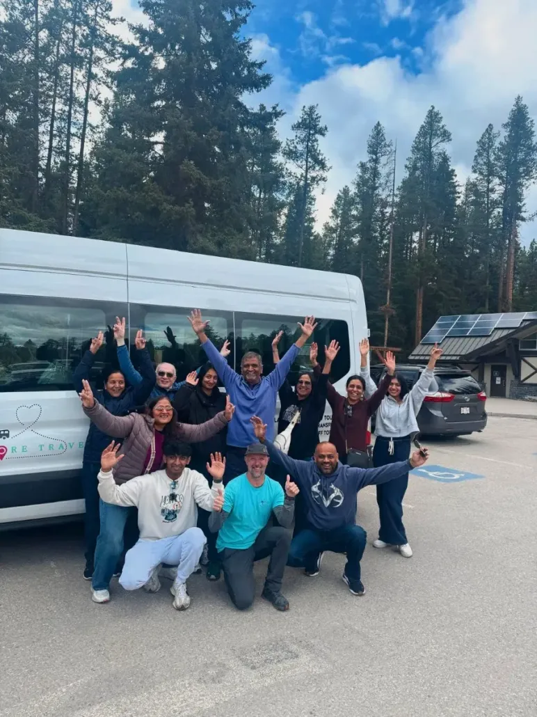 Group of people posing cheerfully in front of a white van in a forested parking area.