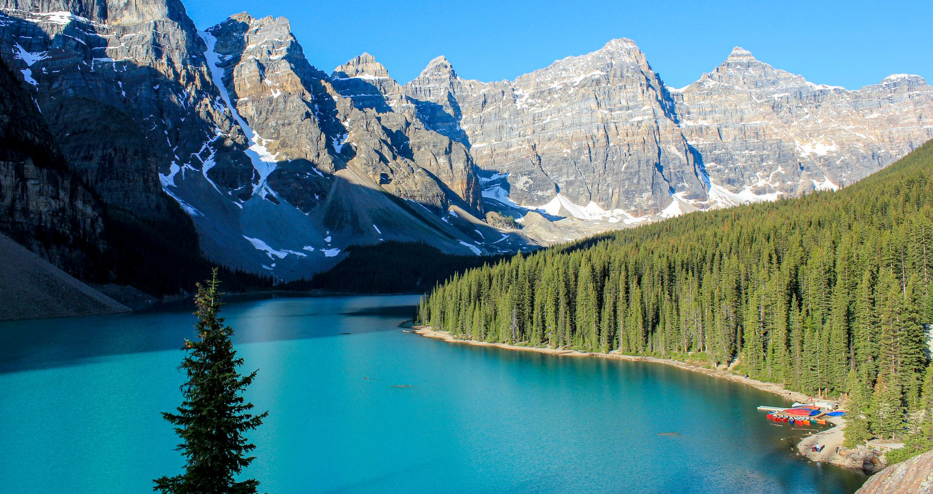 Turquoise alpine lake bordered by pine forest and rugged mountains under a bright blue sky