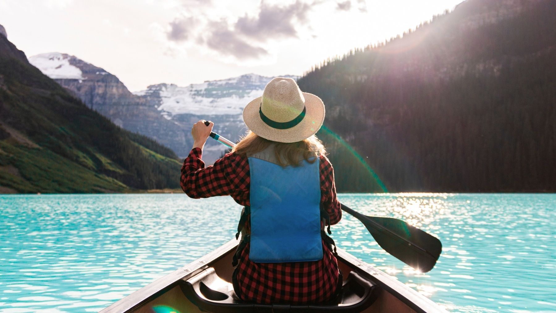 Person canoeing on a turquoise lake with mountains and sunlight in the background