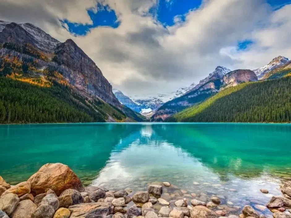 Lake Louise reflecting snowy peaks and cloudy sky, with rocky shoreline in the foreground