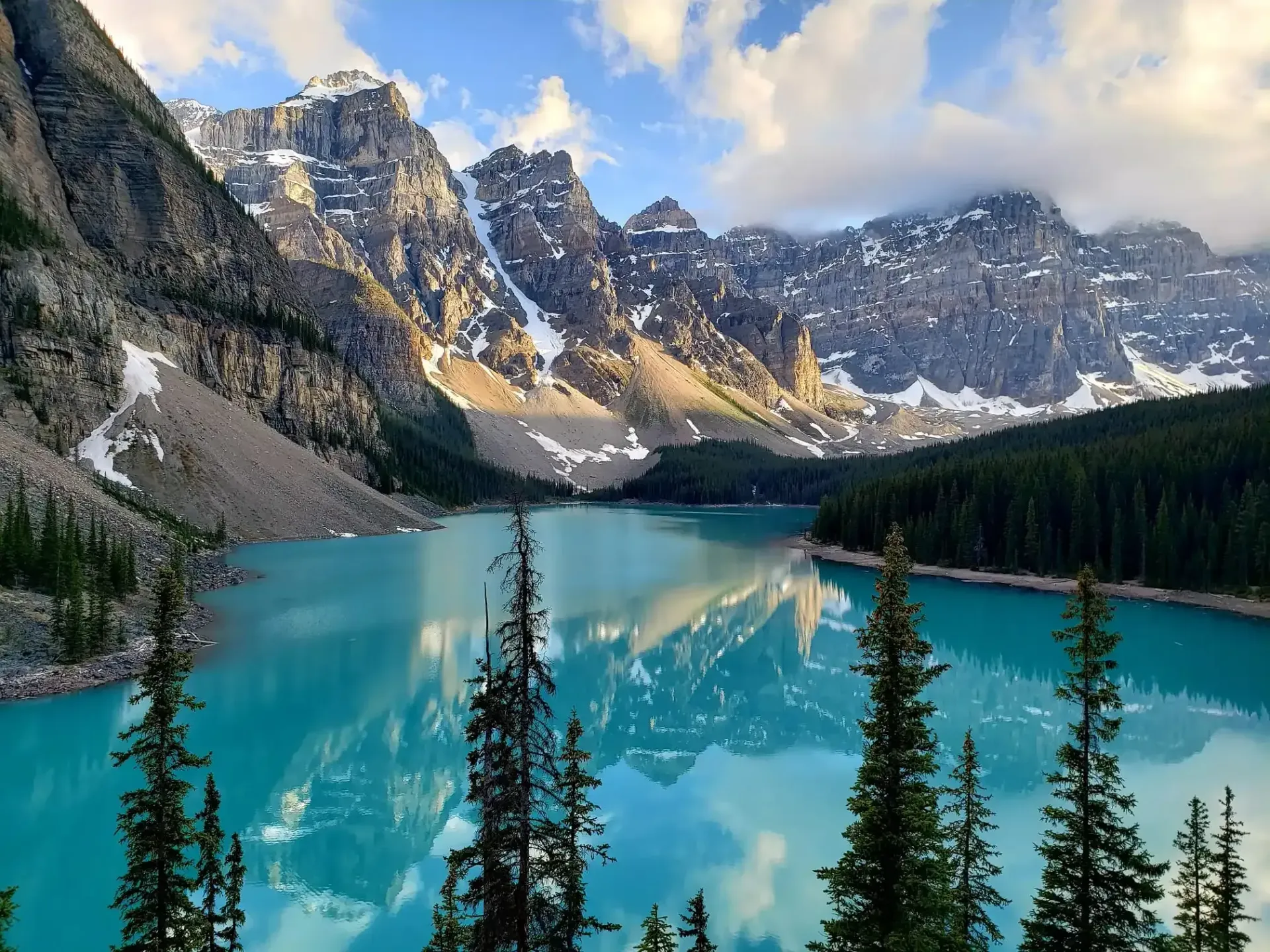 Turquoise alpine lake surrounded by rugged mountains, pine trees, and snow patches under cloudy skies