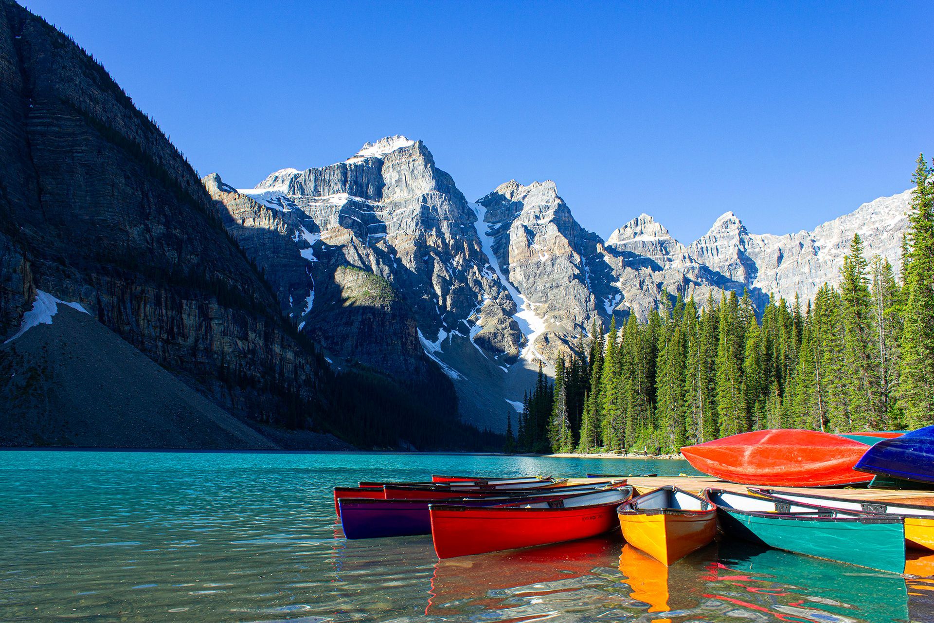 Red canoes on a turquoise mountain lake with pine trees and snow-capped peaks under a blue sky