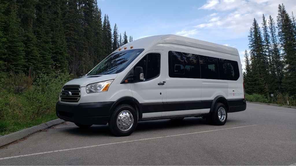 White passenger van parked on a mountain road beside a pine forest under a partly cloudy sky