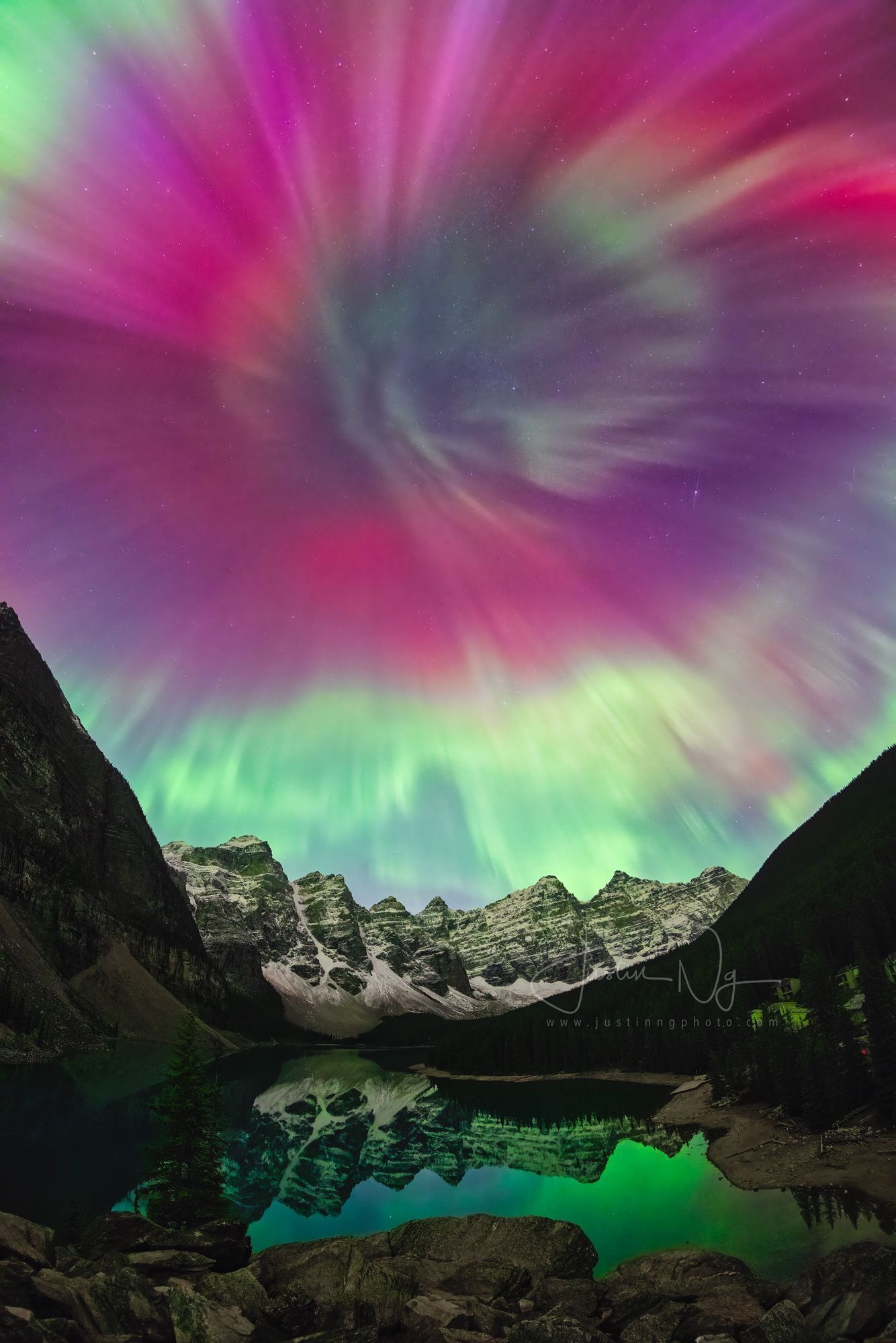 Vibrant pink, green, and purple aurora over snowy mountains reflected in a calm alpine lake