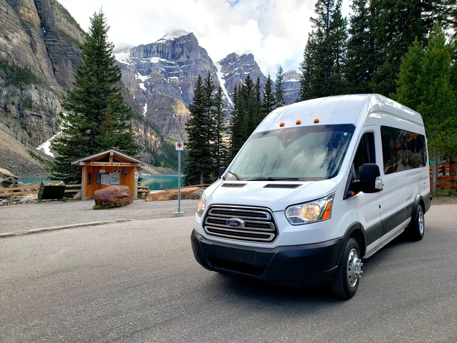 White passenger van parked on a mountain road beside tall pines and rocky cliffs