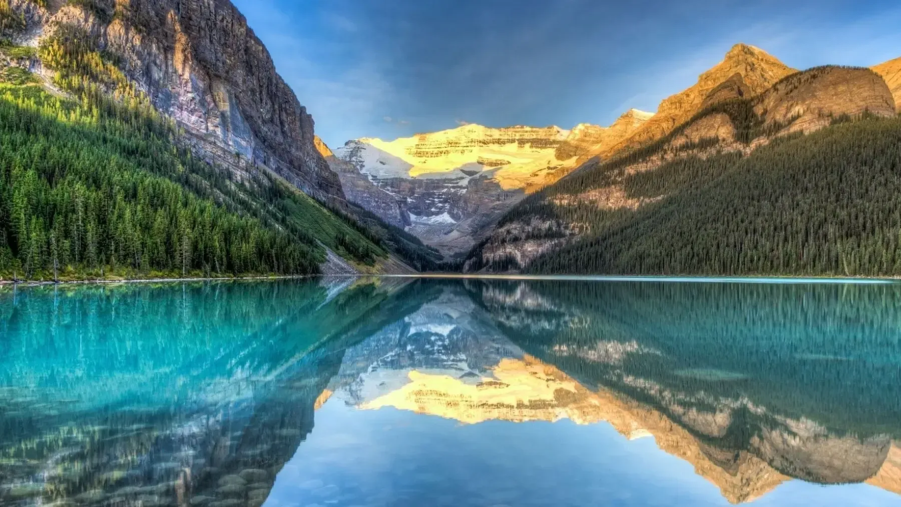 Turquoise alpine lake reflecting forest, mountains, and sunlit peaks under a clear sky