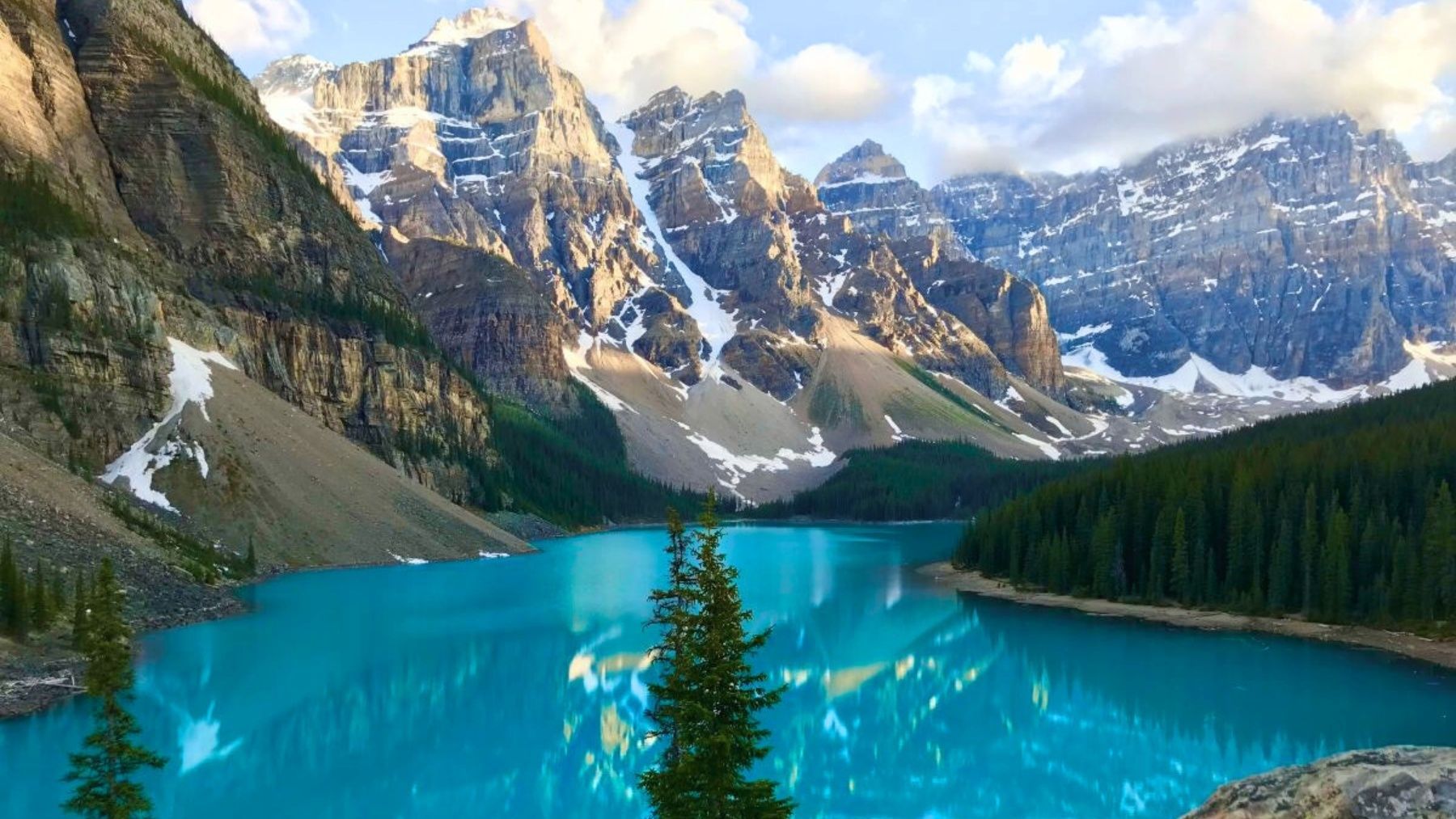 Turquoise mountain lake framed by snowy peaks and pine forest, with a person photographing the view.