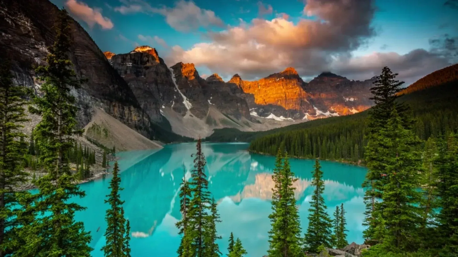 Turquoise mountain lake beneath snow-dusted peaks and orange-lit cliffs at sunset, with pine trees in the foreground